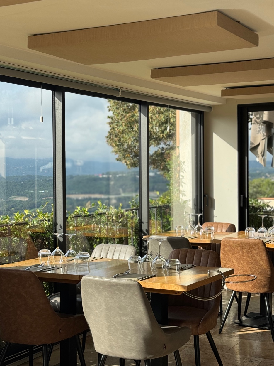 Salle du restaurant aux grandes baies vitrées, tables dressées avec vue sur les collines boisées