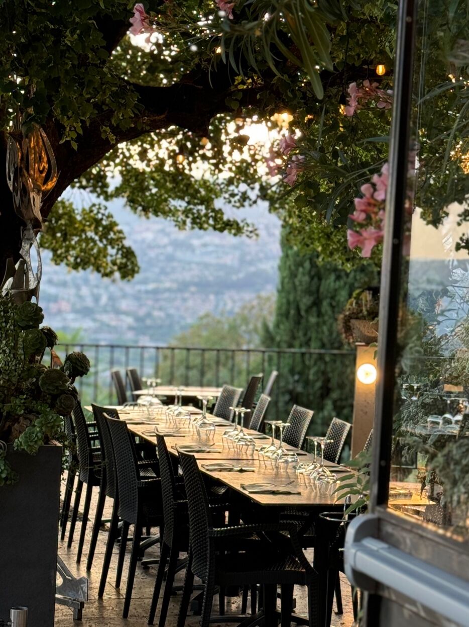 Longue table dressée sous un arbre, guirlandes lumineuses et fleurs roses, vue sur la vallée
