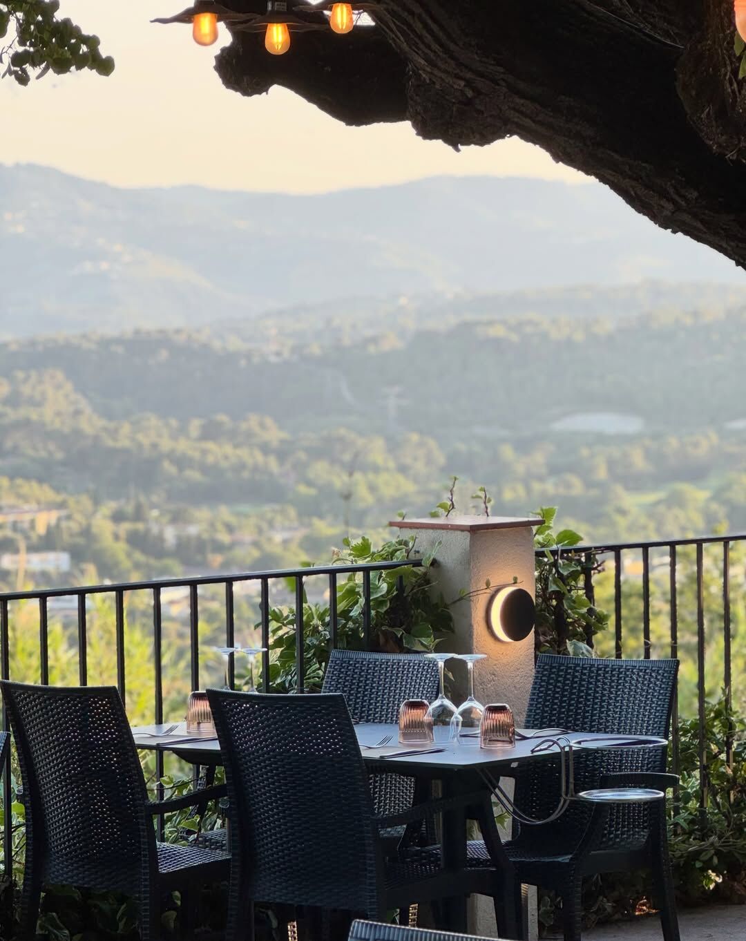 Table et chaises en osier au bord de la terrasse, sous un vieil arbre aux guirlandes lumineuses, au coucher du soleil sur les collines